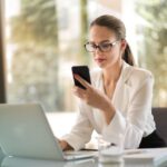 Eine Frau in legerem Business-Outfit schaut vor ihrem Laptop in ein Telefon.