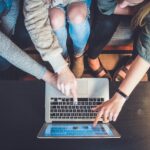 Drei Personen sitzen auf einem Sofa und zeigen auf einen Laptop.
