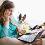 Ein Mädchen mit Kopfhörern, das auf dem Bett sitzt, mit ihrem Laptop und ihrem Hund neben sich.