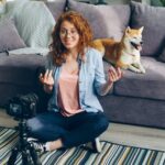 Eine rothaarige Frau lächelt und spricht vor einer Kamera, während ihr Hund hinter ihr steht.