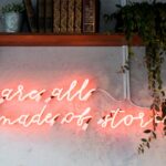 Un cartel de neón rosa en la pared, bajo una estantería, que dice: "Todos estamos hechos de historias".