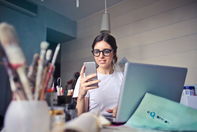 Una mujer mira su teléfono mientras trabaja con un portátil.