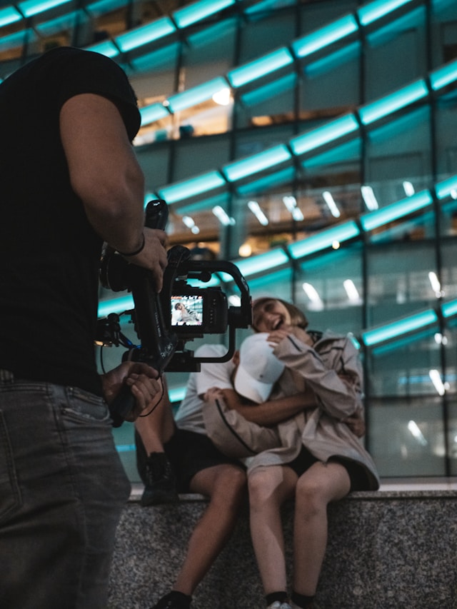Un hombre con camiseta negra filma a una pareja sentada en cemento gris. 
