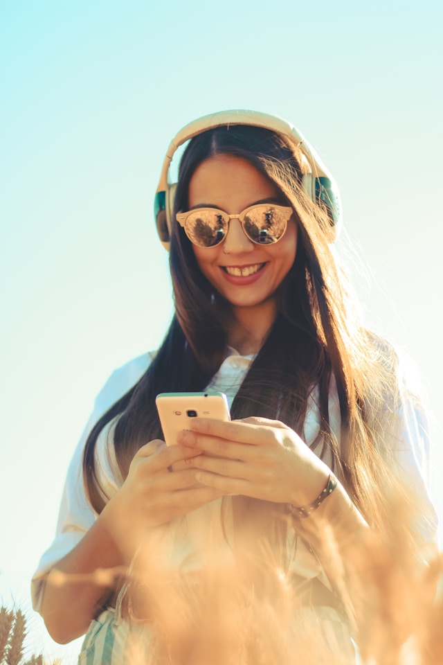 Une femme portant des lunettes de soleil et des écouteurs sourit à son téléphone.