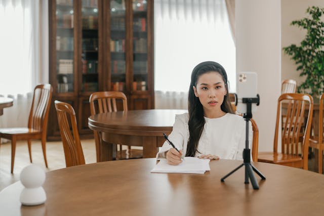 Une femme assise à une table avec un stylo et des papiers tout en faisant face à un téléphone sur un trépied.