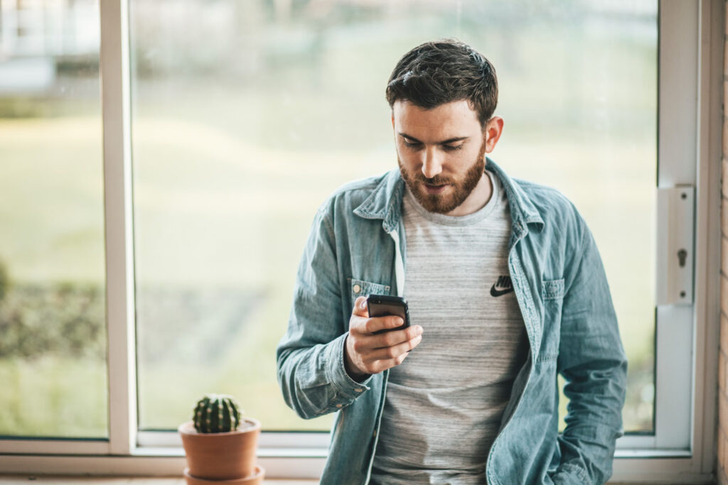 Un homme se tient devant une fenêtre, tenant un téléphone d'une main et le regardant vers le bas.