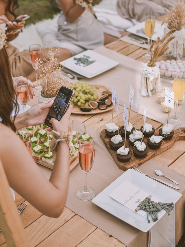 Une table est dressée avec des assiettes décoratives contenant de la nourriture et des boissons. Une femme se penche pour prendre une photo avec son smartphone.