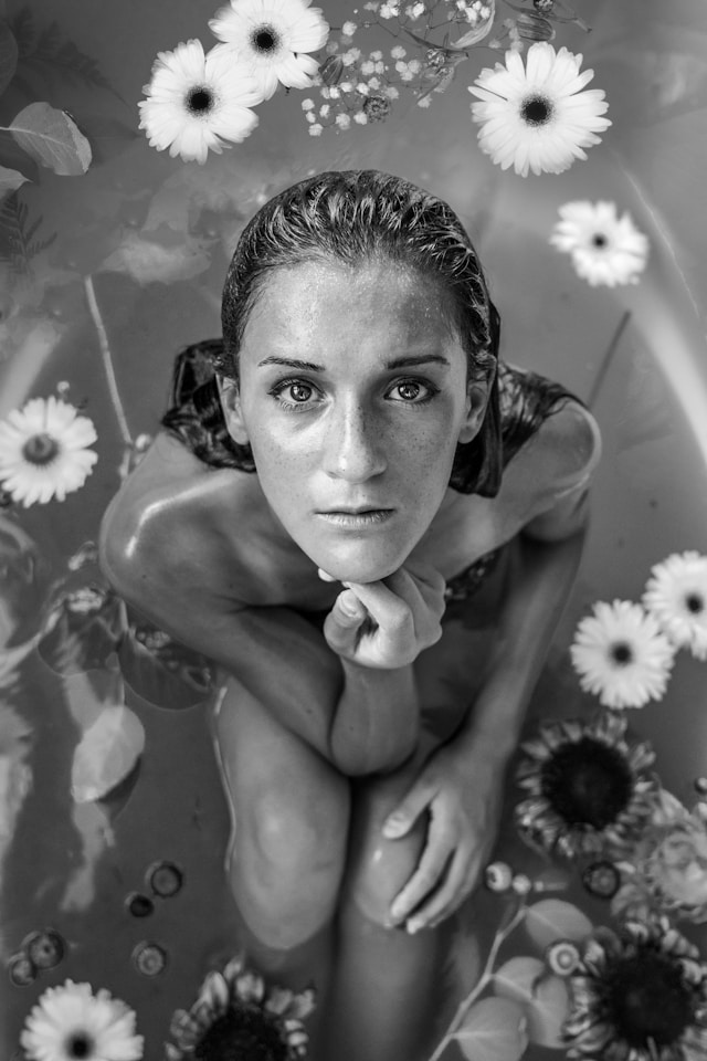 Photo en noir et blanc d'une femme assise dans une bassine d'eau entourée de fleurs.