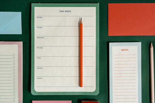 Un calendrier hebdomadaire reposant sur une surface verte et entouré de blocs-notes et de deux crayons.
