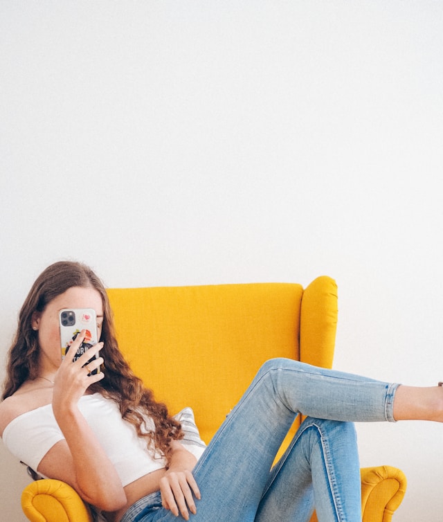 Una donna con un top bianco off-shoulder e jeans blu denim che si fotografa seduta su un divano giallo imbottito.