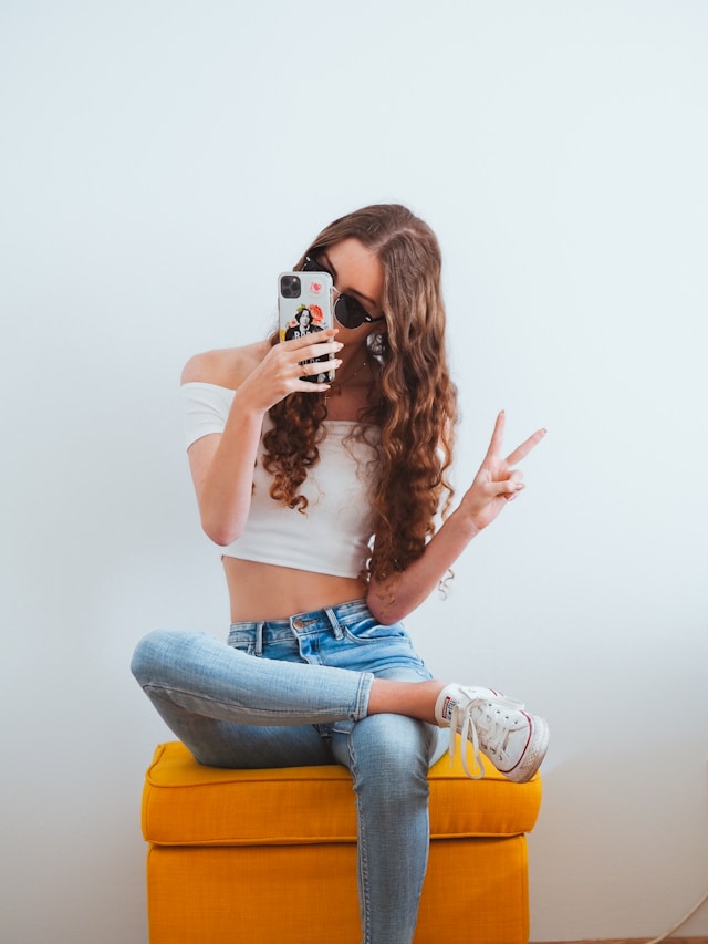 Una donna con un top bianco off-shoulder e jeans blu denim che si fotografa seduta su uno sgabello giallo imbottito.