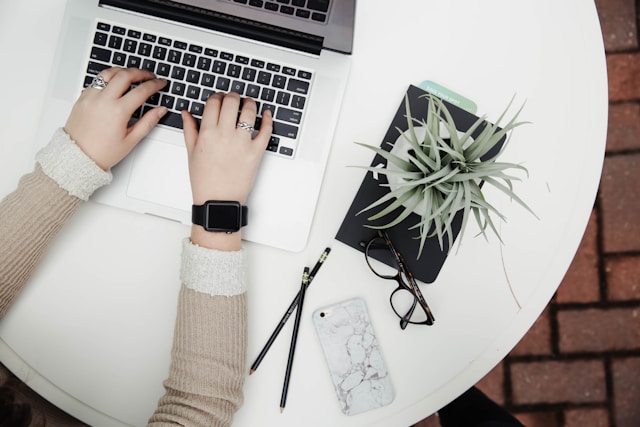 Una persona che utilizza un computer portatile su un tavolo bianco con una pianta di aloe vera, alcuni articoli di cancelleria, un paio di occhiali e un telefono.