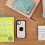 Una scrivania con alcuni quaderni, uno smartphone, una tastiera, un computer portatile e un libro intitolato "Le regole di Instagram".
