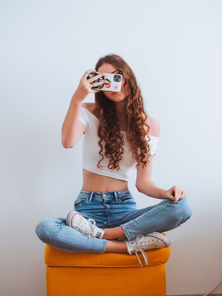 Ragazza seduta su uno sgabello arancione con in mano uno smartphone per scattare una foto. 