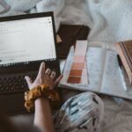 Una persona che digita un'e-mail su un computer portatile mentre è seduta su un letto con accanto una tavolozza di colori e un taccuino.