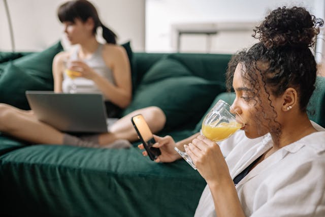 Una donna seduta sul divano di casa mentre beve un succo d'arancia e consulta il suo telefono.