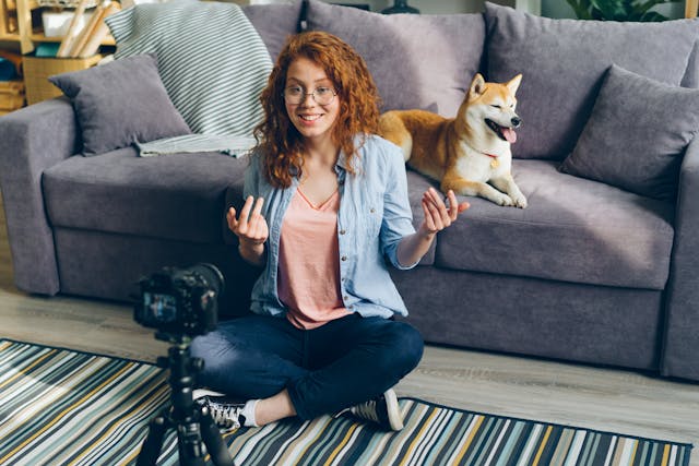 Una donna dai capelli rossi che sorride e parla davanti a una telecamera con il suo cane domestico alle spalle.