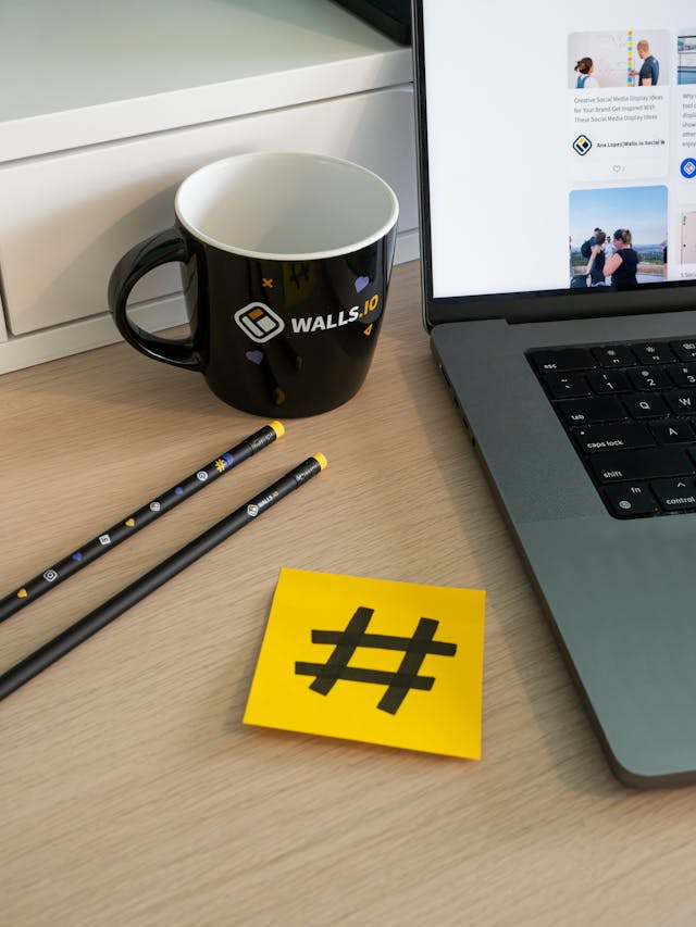 Uma secretária de escritório com um computador portátil, uma caneca de café, alguns artigos de papelaria e uma nota adesiva amarela com uma hashtag desenhada.