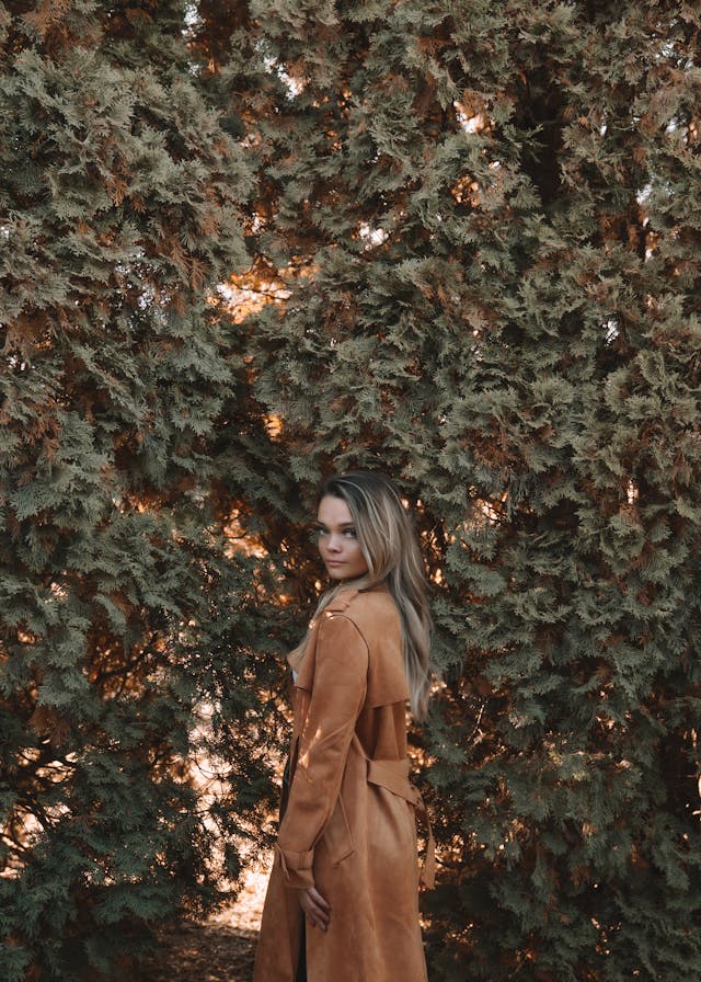 Uma mulher com um casaco castanho, parada em frente a folhas de árvores densas.