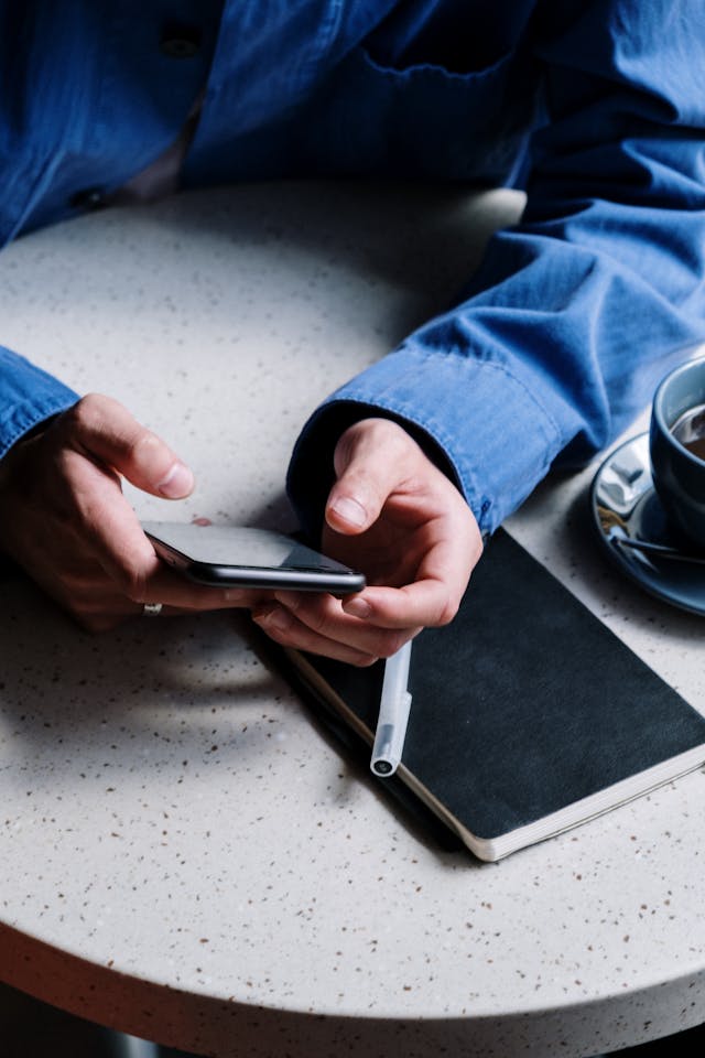 O persoană își ținea telefonul la o masă de teracotă, iar mâinile sale se aflau deasupra unui jurnal și a unui stilou.