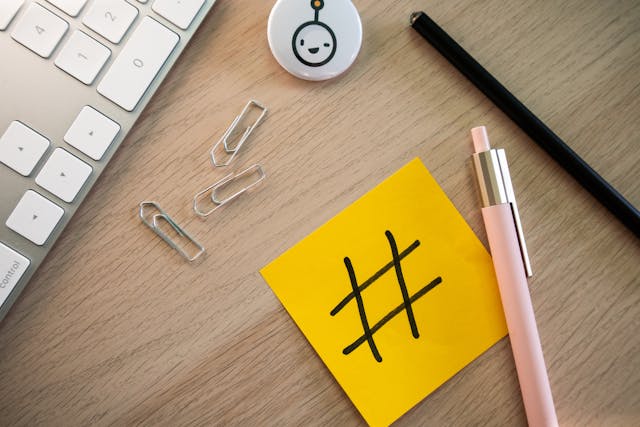 Vedere de sus a unei tastaturi, a unor articole de papetărie și a unui bilet autocolant cu un hashtag pe el, așezate pe un birou.
