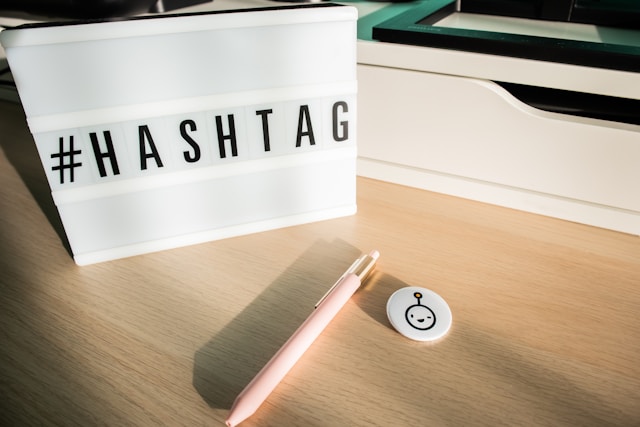 Un birou din lemn cu un pix, o insignă și un semn pe care scrie "#HASHTAG".