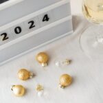 Un semn alb cu un hashtag și cifre care arată 2024 lângă un pahar de șampanie și globuri aurii.
